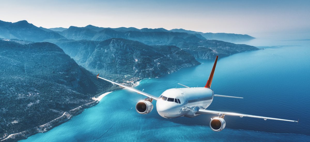 Aircraft is flying over islands and sea at sunrise in summer. Landscape with white passenger airplane, seashore, mountains, forest, clear sky, and blue water in bright day. Travel and resort. Tourism